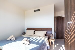 Ein ordentlich bezogenes Bett mit einer blauen Steudecke, zwei Kissen und gerollten Handtüchern. Ein kleiner Nachttisch mit einer Lampe steht neben dem Bett.