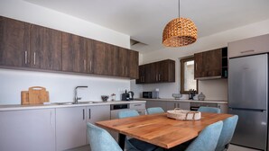 A modern kitchen with dark wooden cabinets, a stainless steel refrigerator, a wooden dining table with blue chairs, and a woven pendant light. - Elpida Luxury Villa, Private Pool, Fantastic View (Chania)