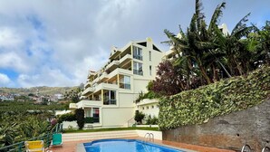 A swimming pool with a tiled deck, a view of a hillside residential area, and a building with balconies. - Apartamento Precioso | Large terrace, sea view and pool (Arco da Calheta)