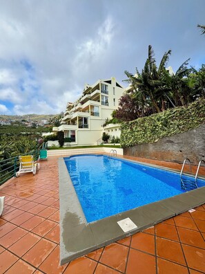 A swimming pool with a tiled deck, a view of a hillside residential area, and a building with balconies. - Apartamento Precioso | Large terrace, sea view and pool (Arco da Calheta)