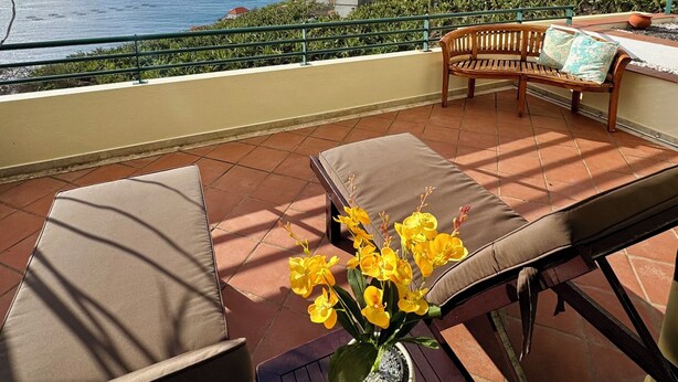 A terrace with a view of the sea, a wooden bench, a table with a potted plant, and a railing. - Apartamento Precioso | Large terrace, sea view and pool (Arco da Calheta)
