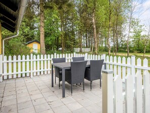 An outdoor patio with a table and chairs, surrounded by a white picket fence, set against a backdrop of trees and a yellow building. - 4 Star Holiday Home in Bording (Bording)