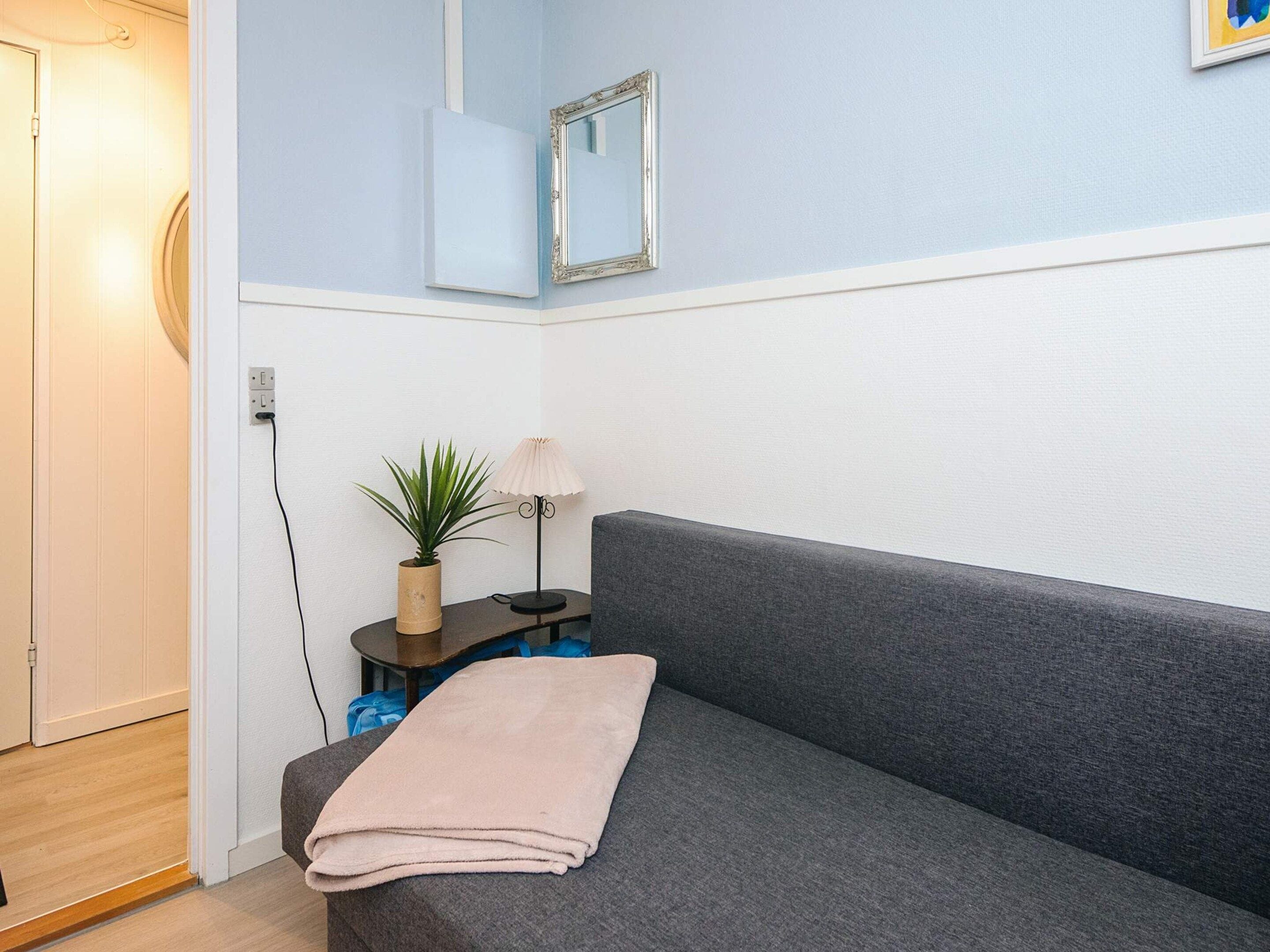 Una habitación con un sofá gris, una mesita con una lámpara y una planta, un espejo adosado a la pared y una puerta.