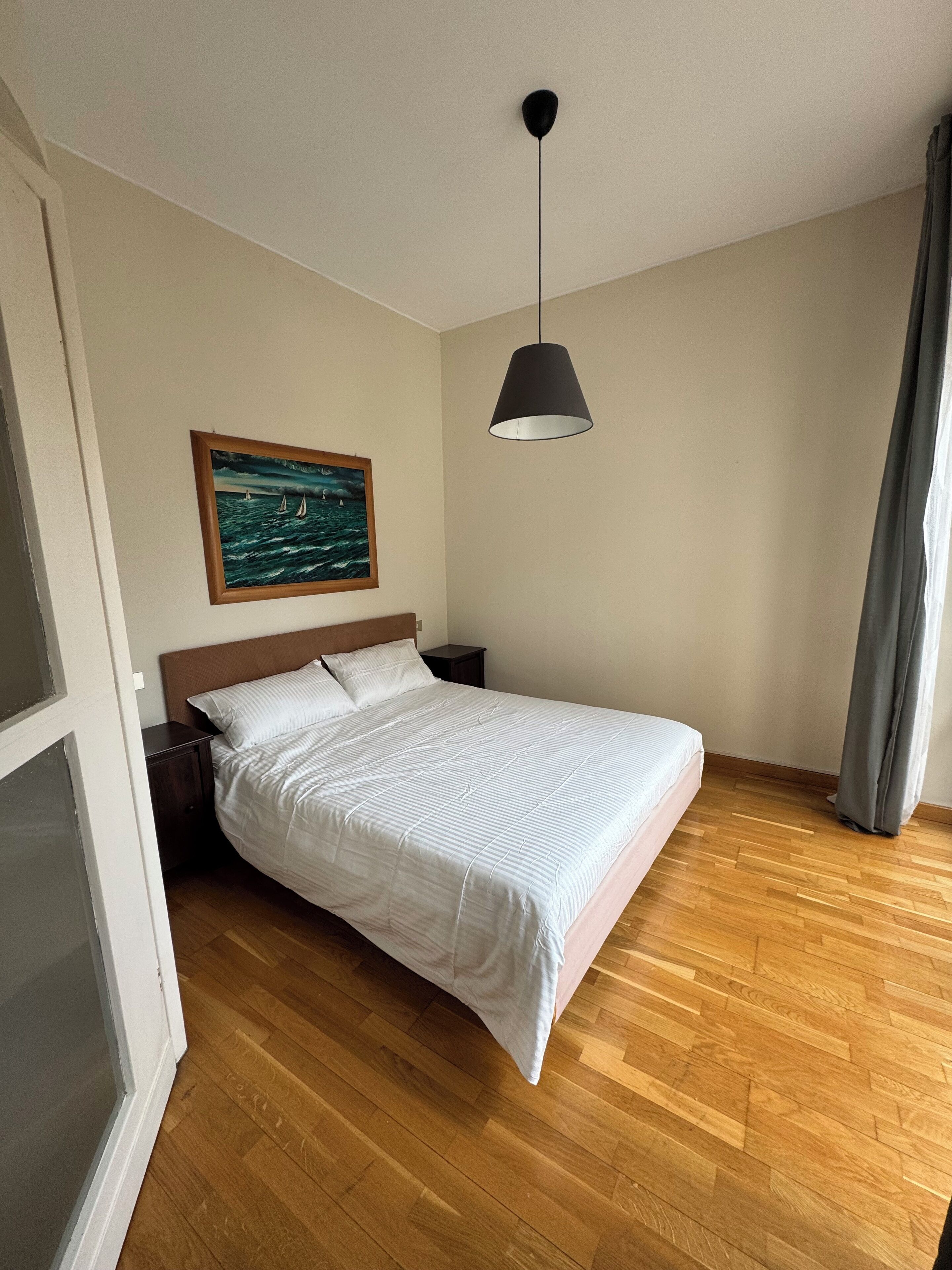 Una camera da letto con un letto, pavimento in legno, un comodino, un quadro appeso al muro e una lampada a sospensione.