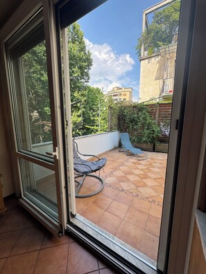 Una porta a vetri scorrevoli che dà su un patio con pavimento piastrellato, due poltrone e vista su un edificio con alberi e cielo sereno.