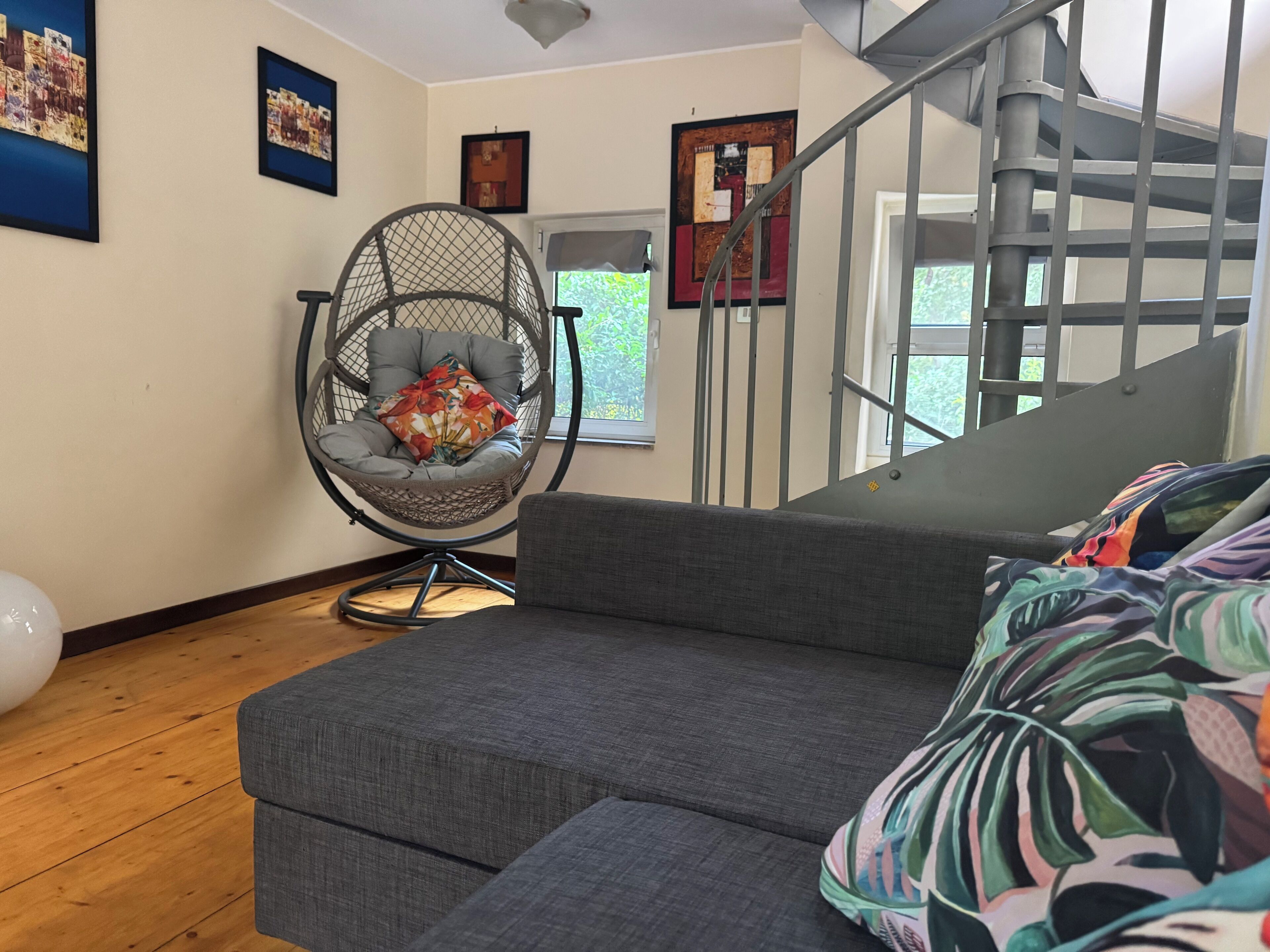 Ein modernes Wohnzimmer mit einem grauen Sofa, einem Hängesessel mit einem floralen Kissen und einer Treppe mit Metallgeländer.
