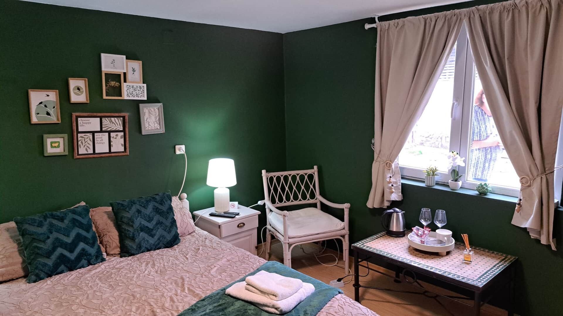 Un dormitorio con cama, una silla, una mesita de noche con lámpara, una mesa con bandeja y una ventana con cortinas.