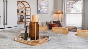 Cuina moderna amb taulell de marbre, una ampolla gran marró i una safata amb objectes de cuina. Hi ha una zona de menjador amb taula i cadires, i un sofà amb coixins. Un lampadari i un quadre adornen la paret.