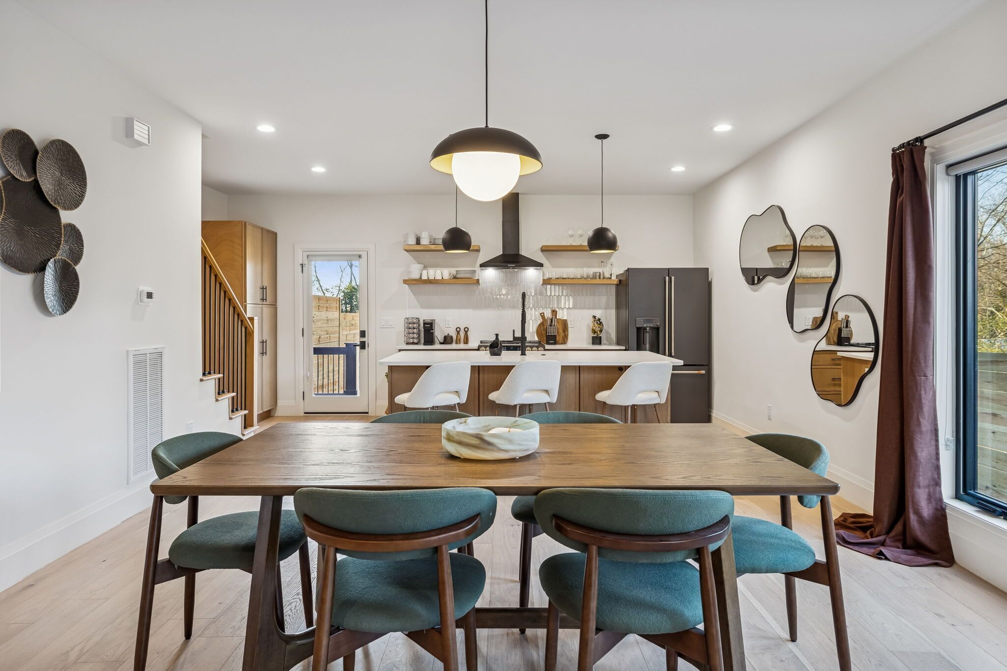Un comedor moderno con una mesa de madera y sillas color turquesa, una cocina al fondo y un ventanal con cortinas.