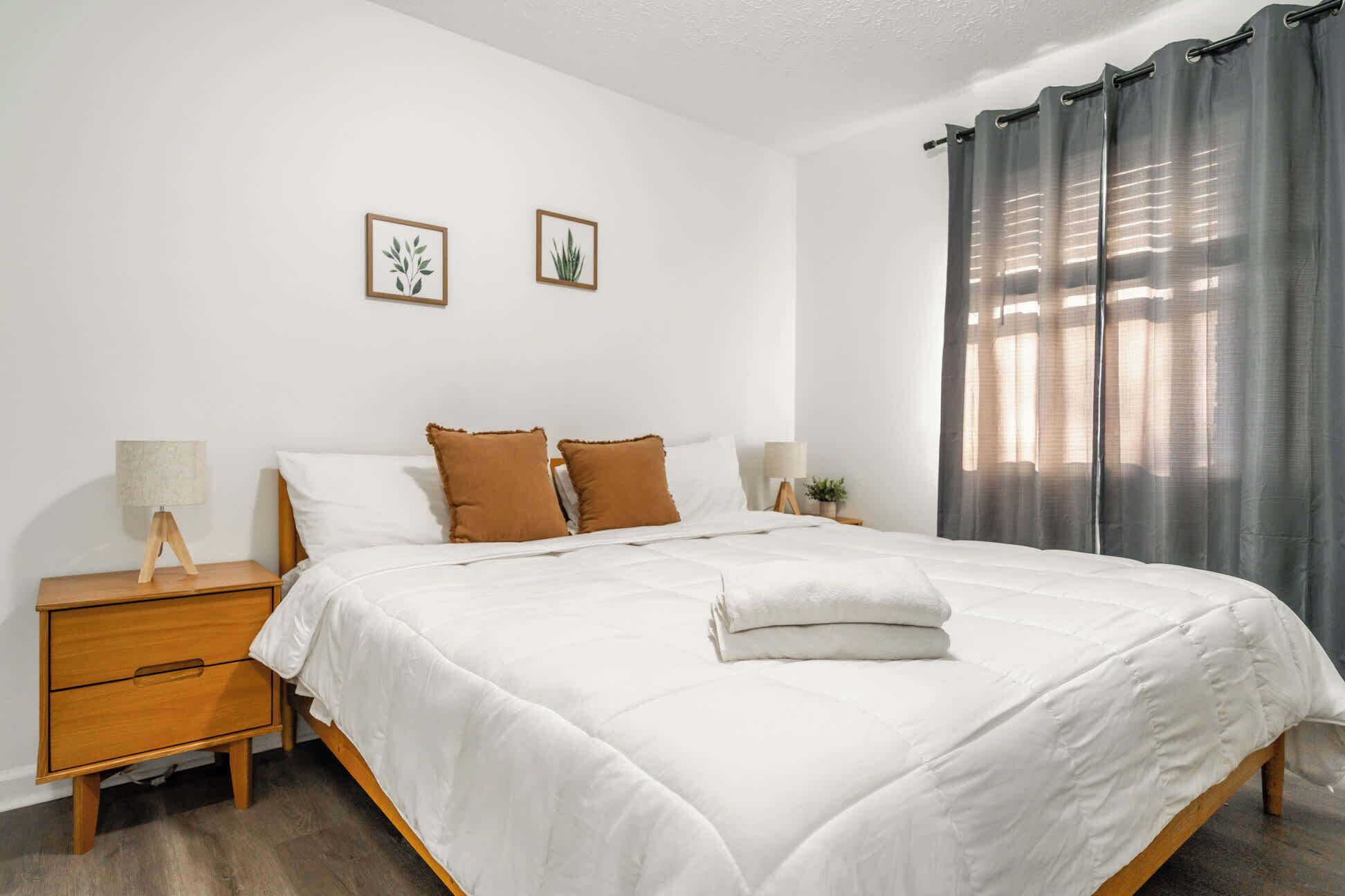 Ein Schlafzimmer mit einem Bett, zwei Nachttischen, einer Lampe, einem Fenster mit Vorhängen und zwei gerahmten Bildern an der Wand.