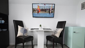 A modern room with a white table, two black chairs, a small plant, and a TV displaying a beach scene. - The Lodge II - with private garden and beach chair (Büsum)