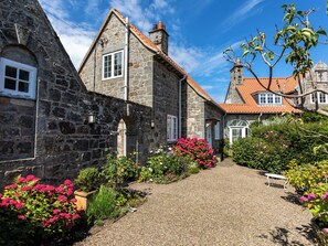 A stone building with a gravel path, surrounded by flowering plants and a bench. - Mid Cote (Seahouses)