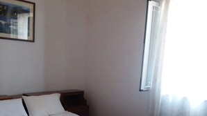Una habitación de hotel con una cama de madera, ropa de cama blanca, una mesita de noche y una ventana con cortinas traslúcidas.