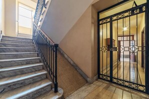 Eine Marmortreppe mit schwarzem Metallgeländer führt zu einem Raum mit einem gemusterten Boden und einem verzierten, schwarzen Tor.
