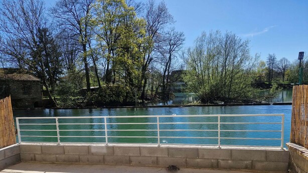 Una terraza de hormigón con barandilla blanca da a un río, rodeada de árboles y un muro de piedra.
