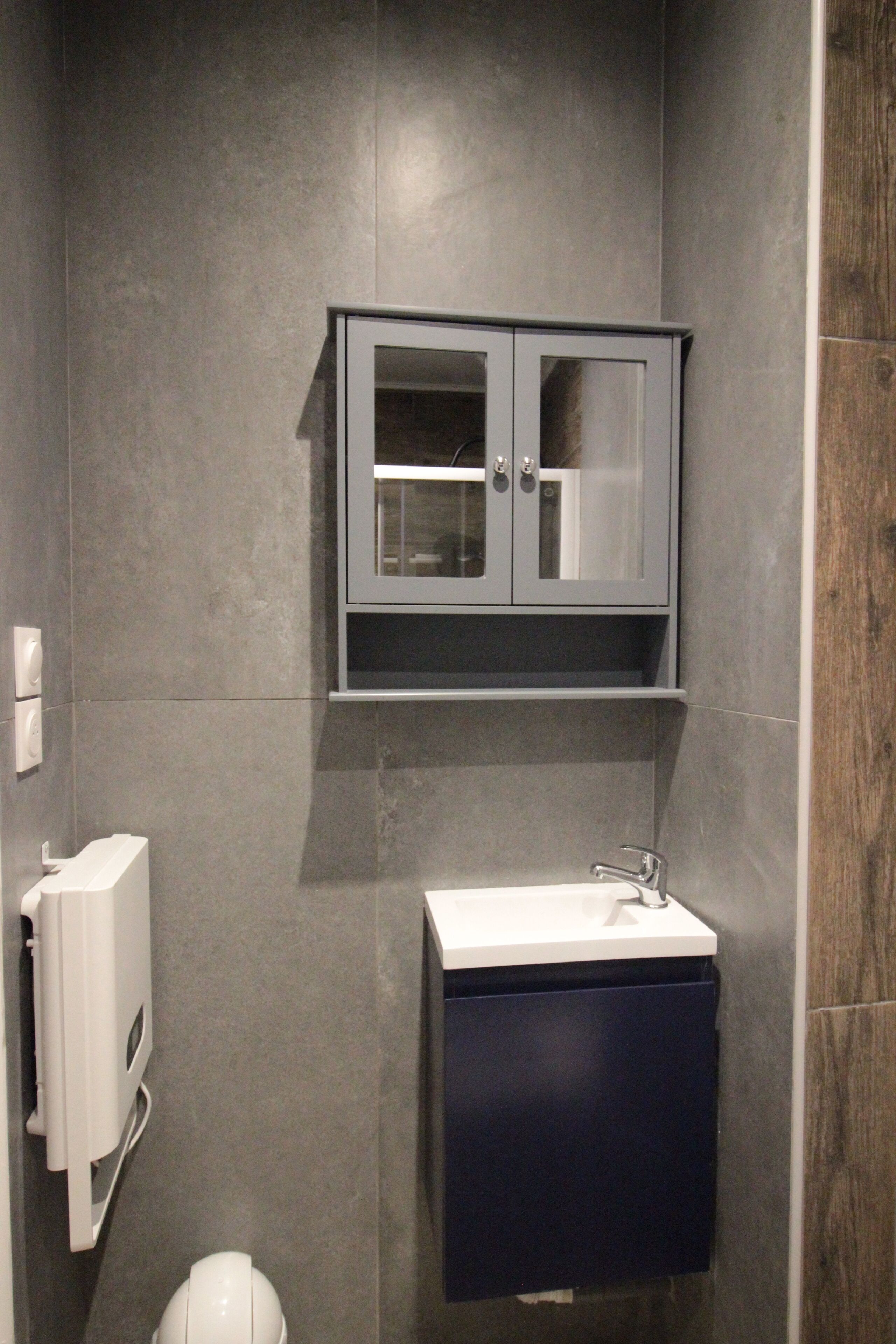 Une salle de bain avec un mur recouvert de carreaux gris, des toilettes blanches, un lavabo blanc avec un meuble sombre en dessous et un meuble gris au-dessus du lavabo.