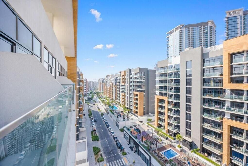 Un edificio moderno de gran altura con varios balcones, cielo despejado y una calle con coches y vegetación.