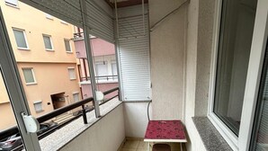 Un petit balcon avec un sol carrelé, une table recouverte d’une nappe rouge et une fenêtre avec des stores.