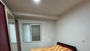 Une chambre à coucher avec un lit, une fenêtre avec des stores, un miroir et un parquet en bois disposé en motif de chevron.