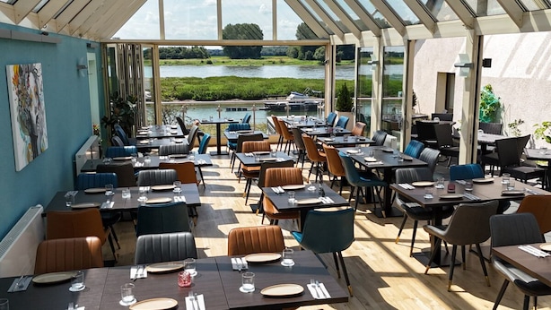An outdoor dining area with tables and chairs, a view of a lake, and a glass roof structure. - Hotel am Hafen Leverkusen (Leverkusen)