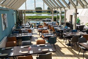 An outdoor dining area with tables and chairs, a view of a lake, and a glass roof structure. - Hotel am Hafen Leverkusen (Leverkusen)