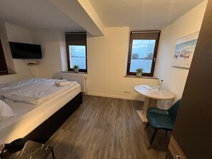 Ein Schlafzimmer mit einem Bett, einem kleinen Tisch mit Stuhl, einem Heizkörper und zwei Fenstern.