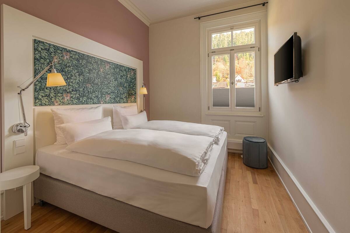 A bedroom with a bed, bedside lamps, a window, and a TV on the wall.
