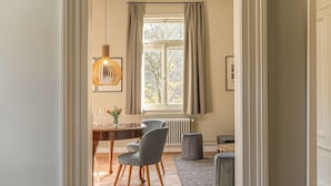 A hallway leading to a dining area with a wooden floor, a table with chairs, a hanging light fixture, and a window with curtains.