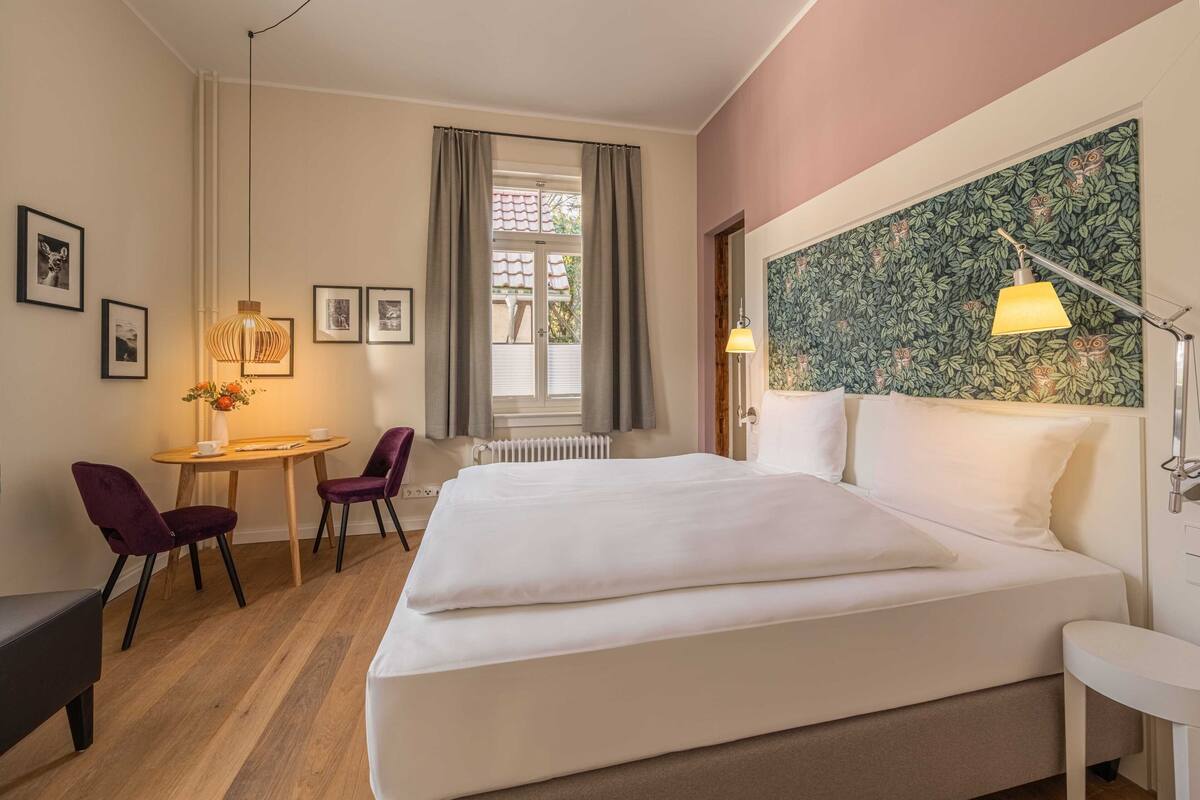A hotel room with a large bed, a small table with chairs, a desk, and a window with curtains.