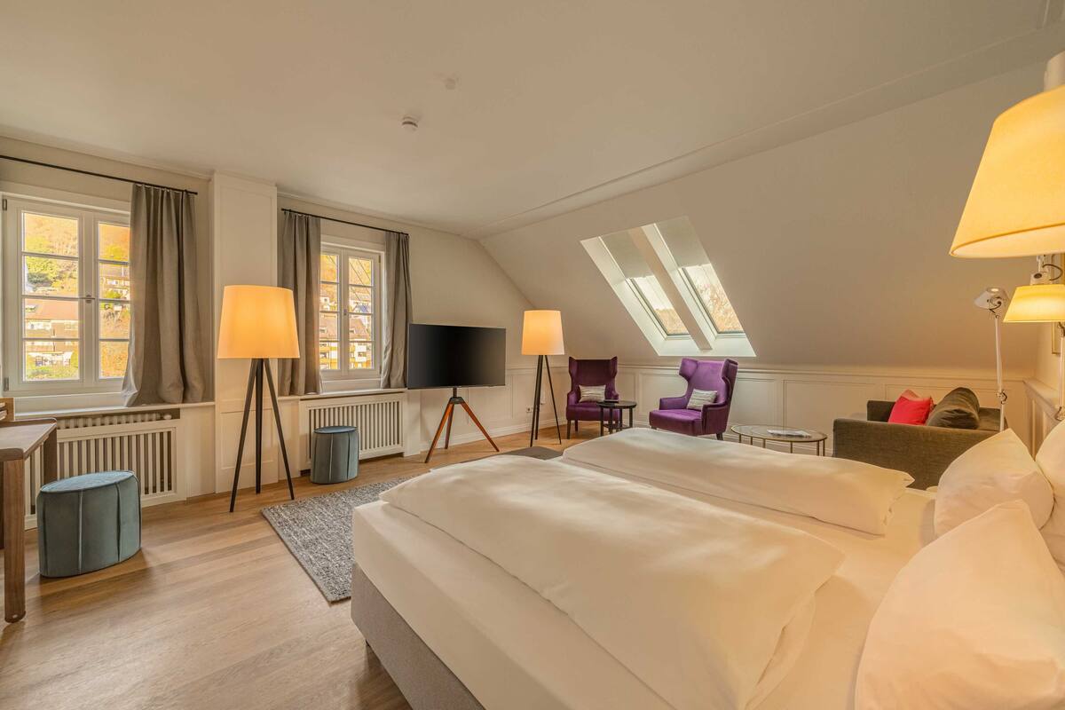 A spacious bedroom with a large bed, two standing lamps, a flat-screen TV, and a skylight.