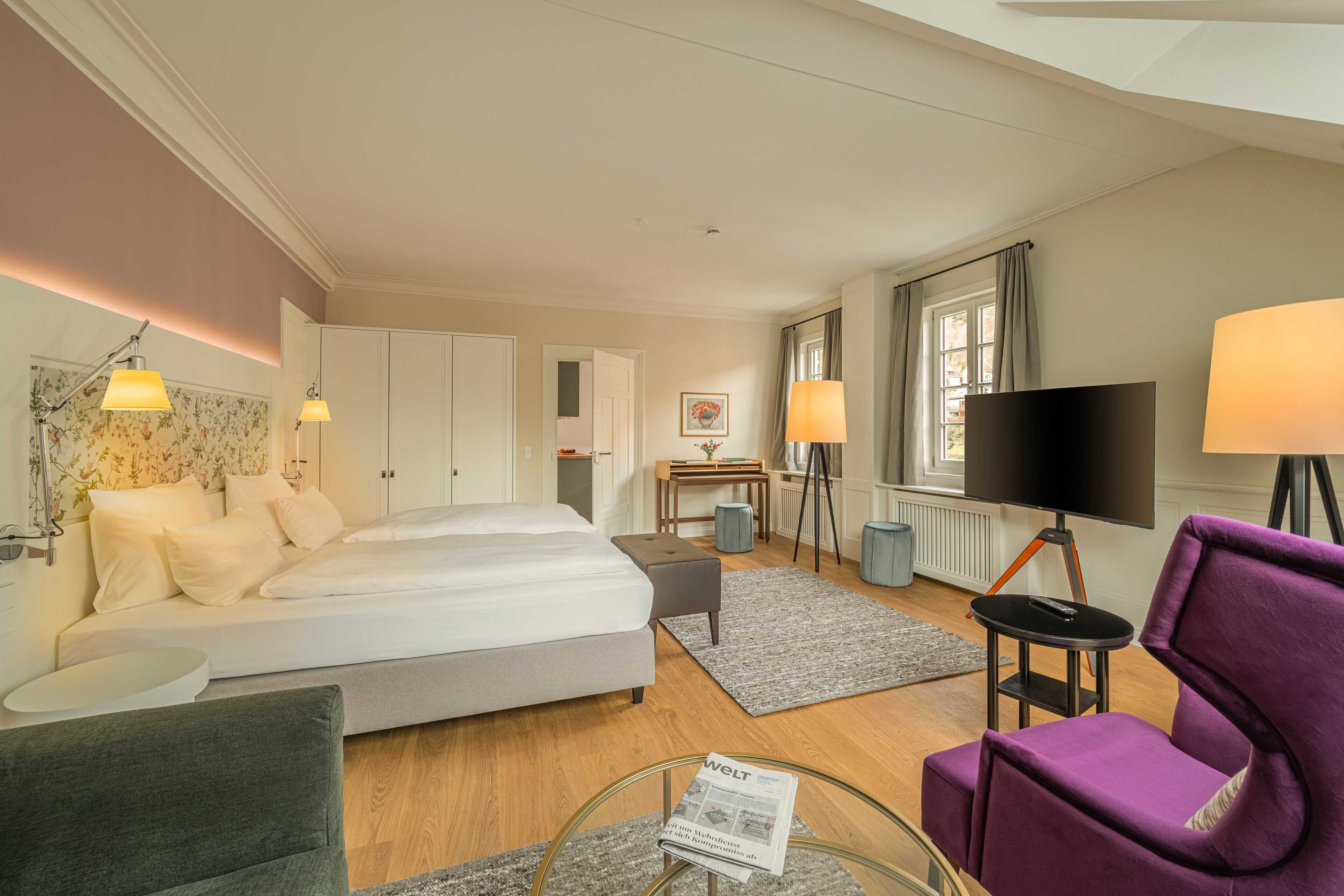 Een hotelkamer met een bed, een bureau, een televisie, een paarse stoel en een glazen salontafel.