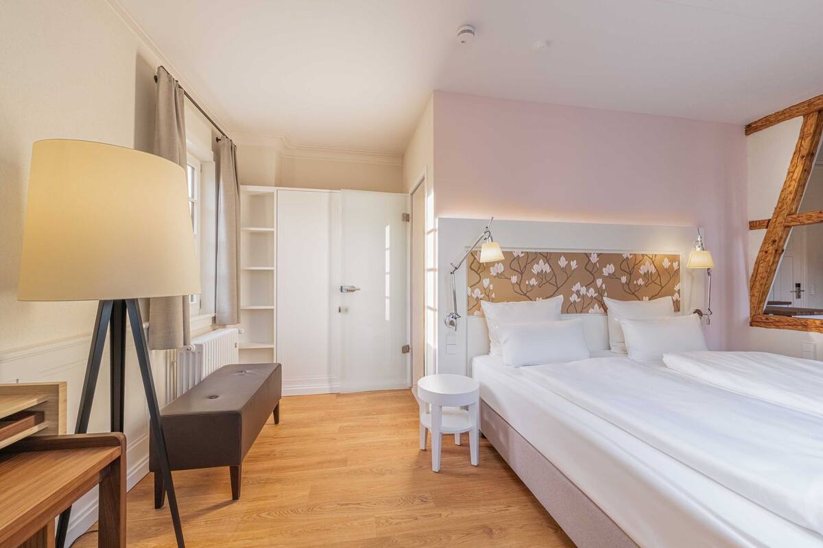 A hotel room with a bed, bedside table, lamp, bench, and a view of the hallway.