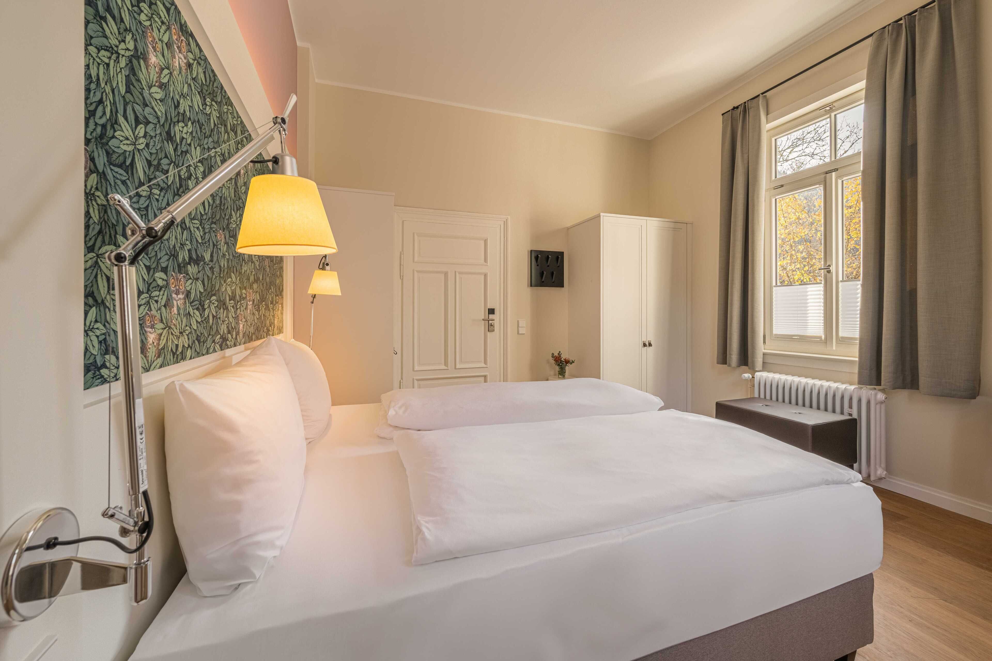 Habitación de hotel con cama, lámparas de noche, radiador, armario y ventana con cortinas.