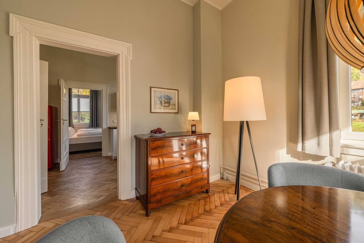 A room with a wooden herringbone floor, a wooden dresser with drawers, a round table, and a lamp.