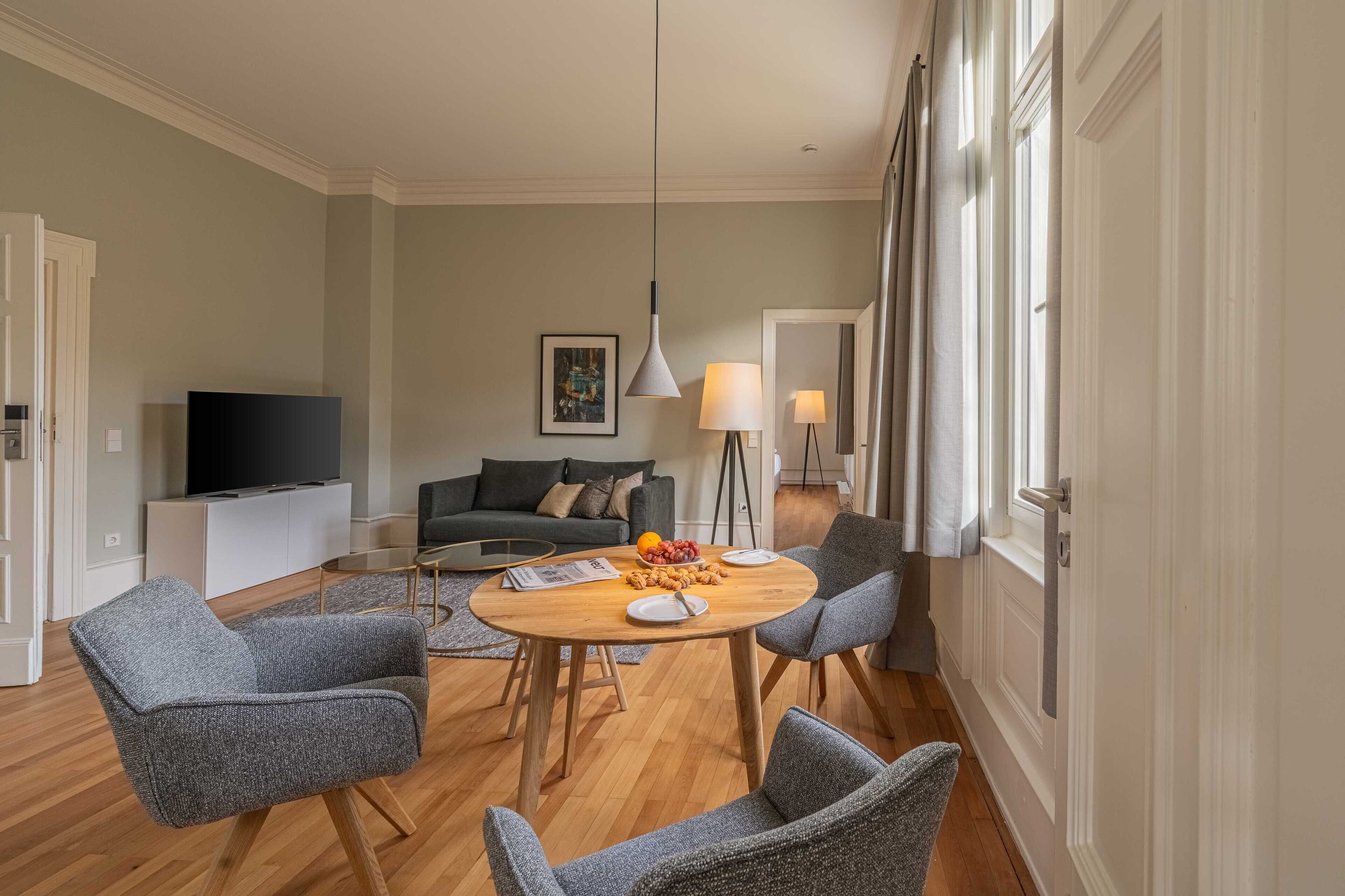 Een moderne woonkamer met een ronde houten tafel, gestoffeerde grijze fauteuils, een flatscreen televisie en een bank.