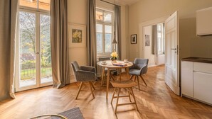 A spacious room with a wooden herringbone floor, a round dining table with chairs, a kitchen area, and large windows with curtains.