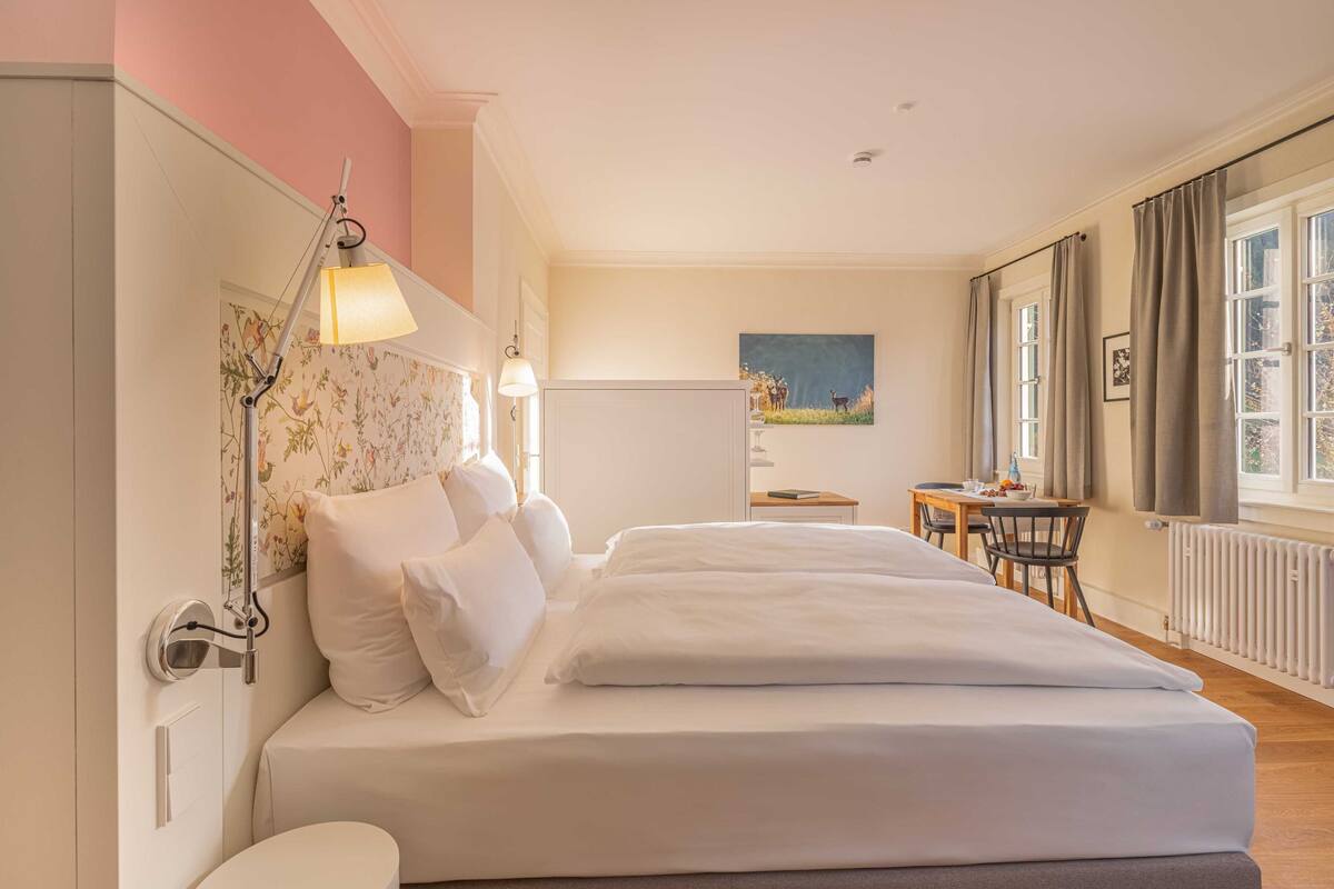 A hotel room with a large bed, a desk, and a chair. There is a window with curtains and a painting on the wall.