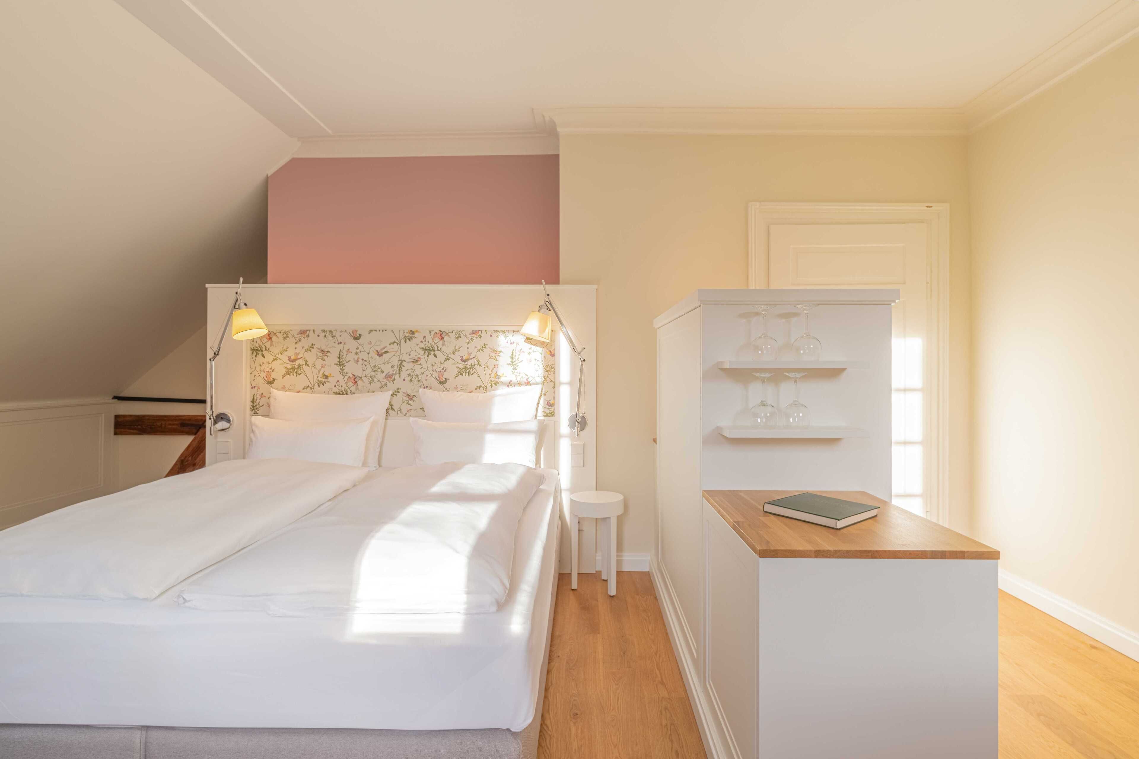 A bedroom with a bed, bedside lamps, a small white table, and a built-in shelf with glasses.
