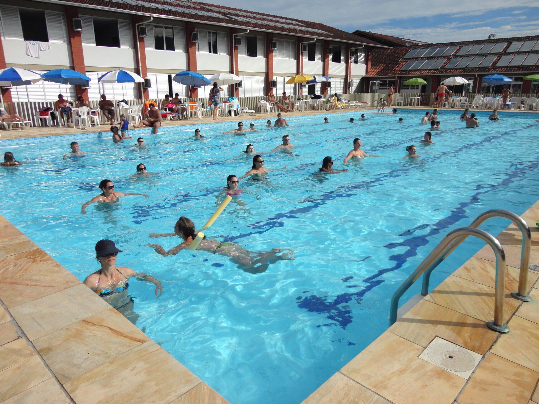 A swimming pool with people swimming and lounging. There are umbrellas and lounge chairs around the pool.
