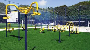 Uma área de exercícios ao ar livre com diversos equipamentos de ginástica, cercada por uma grade azul e um gramado.