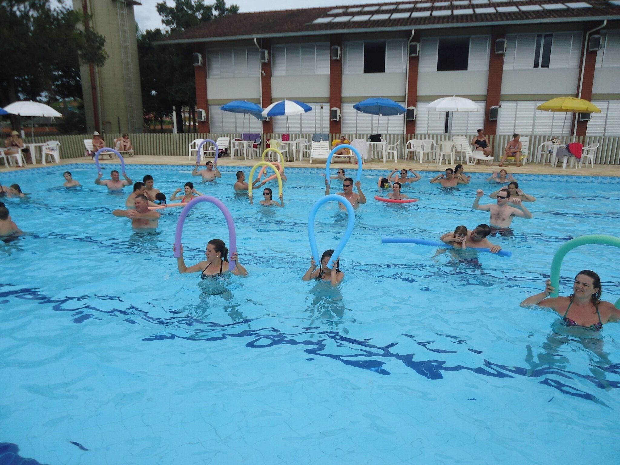 A swimming pool with people using pool noodles. There are umbrellas and lounge chairs around the pool.