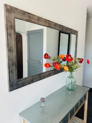 Ein Spiegel mit dunklem Rahmen, eine Vase mit roten und orangefarbenen Blumen sowie eine helle Arbeitsplatte mit einem kleinen Dekoartikel.