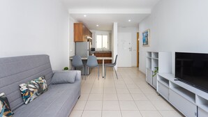 Een moderne woonkamer met een grijze bank, een flatscreen televisie en een eethoek met stoelen, met een keuken op de achtergrond.