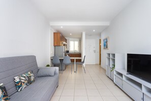 Ein modernes Wohnzimmer mit grauem Sofa, Flachbildfernseher und einem Essbereich mit Stühlen. Im Hintergrund ist eine Küche zu sehen.