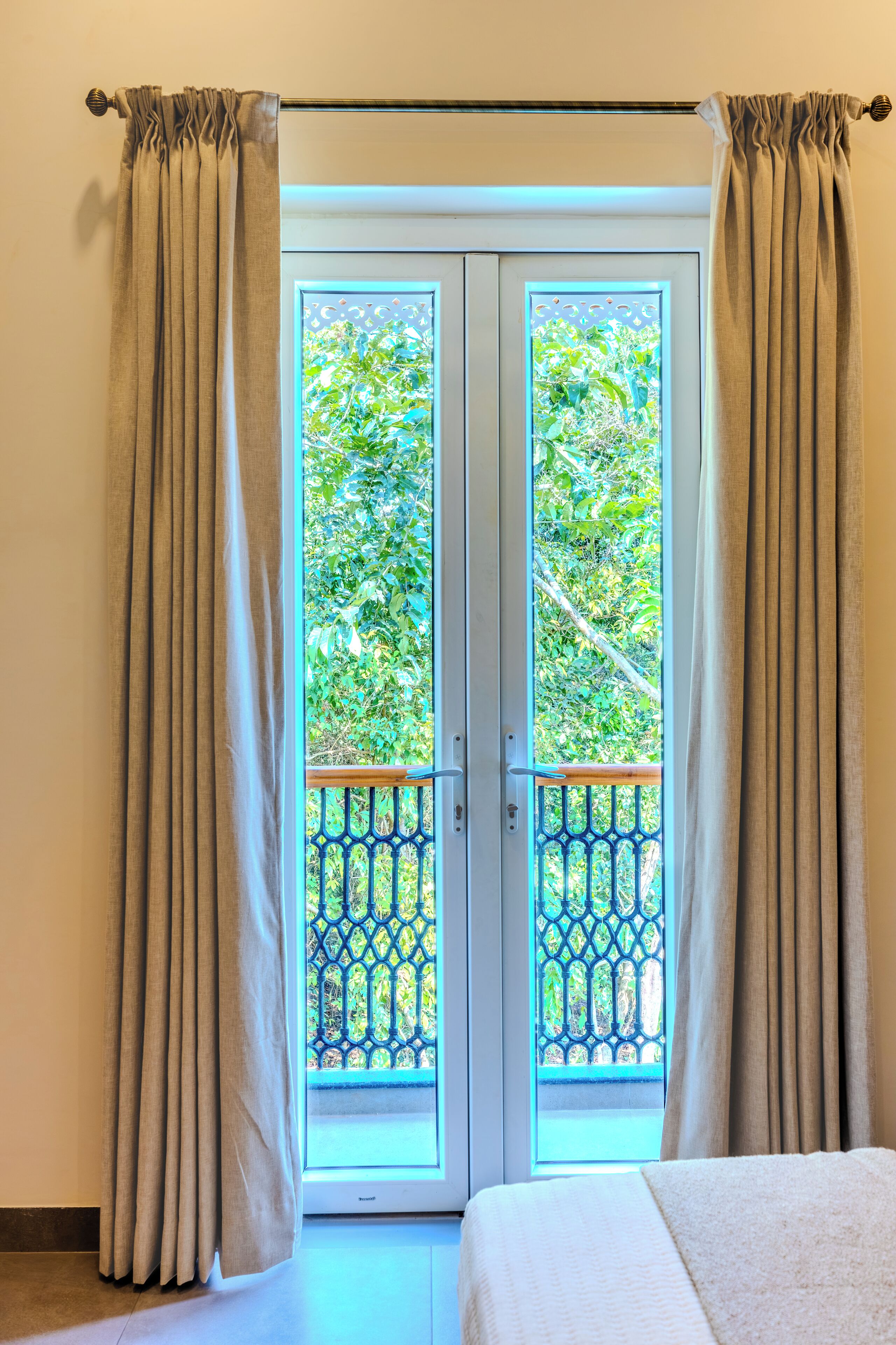 A room with a bed, beige curtains, and a window with a view of greenery.