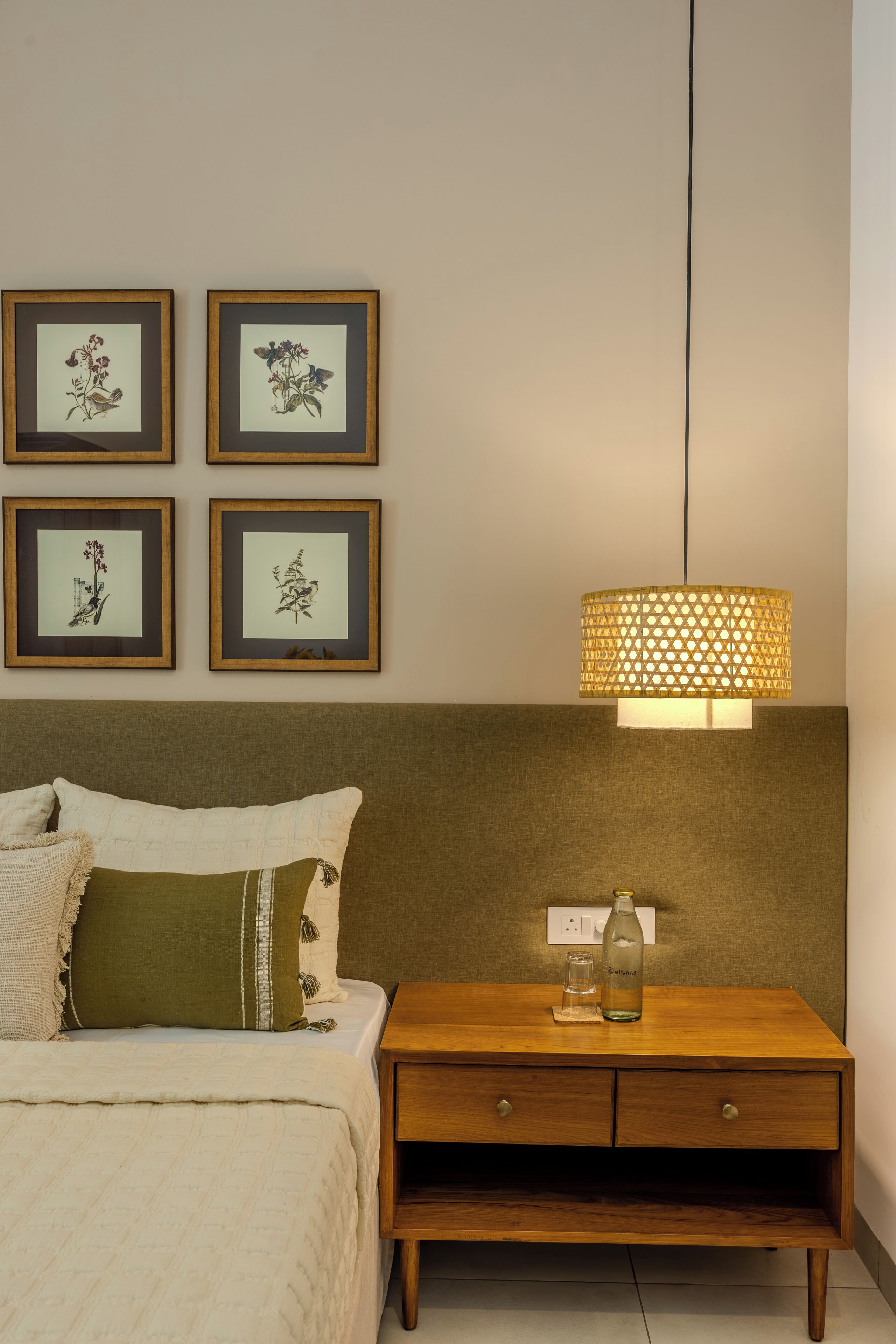 Une chambre à coucher comprenant un lit, une table de chevet, une lampe et quatre reproductions botaniques encadrées accrochées au mur.