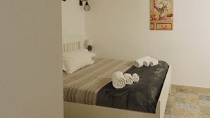 En snyggt bäddad säng med vita kuddar och hoprullade handdukar, ett sängbord med en lampa och ett konstverk som hänger på väggen.