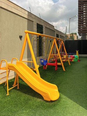 A playground with a yellow slide and swings, set against a building with a concrete wall and a street lamp. - Snug Studio Haven (Accra)