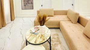 A modern living room with a beige sectional sofa, a round coffee table with a vase and books, a marble floor, and a framed artwork on the wall. - LinQues & Bethel Residences (Ibadan)