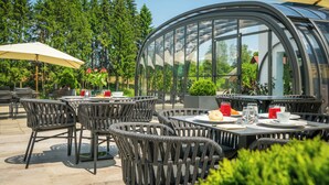 Un patio al aire libre con sillas y mesas de mimbre, una estructura de invernadero de vidrio y vistas a árboles y un edificio al fondo.